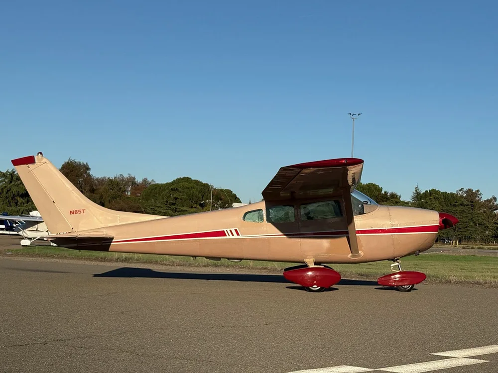 A beige and red Cessna 182 aircraft, registration N85T, parked on an airfield with trees in the background.