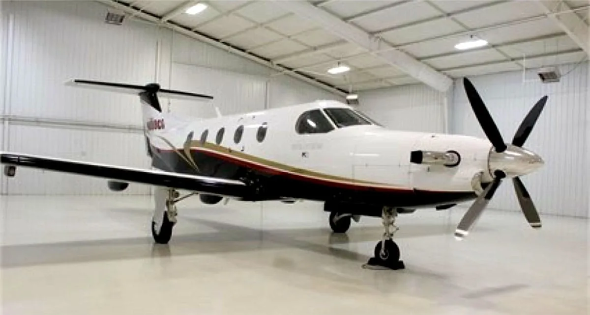 A front-quarter view of a white Pilatus PC-12 turboprop aircraft with black and gold trim inside a hangar.