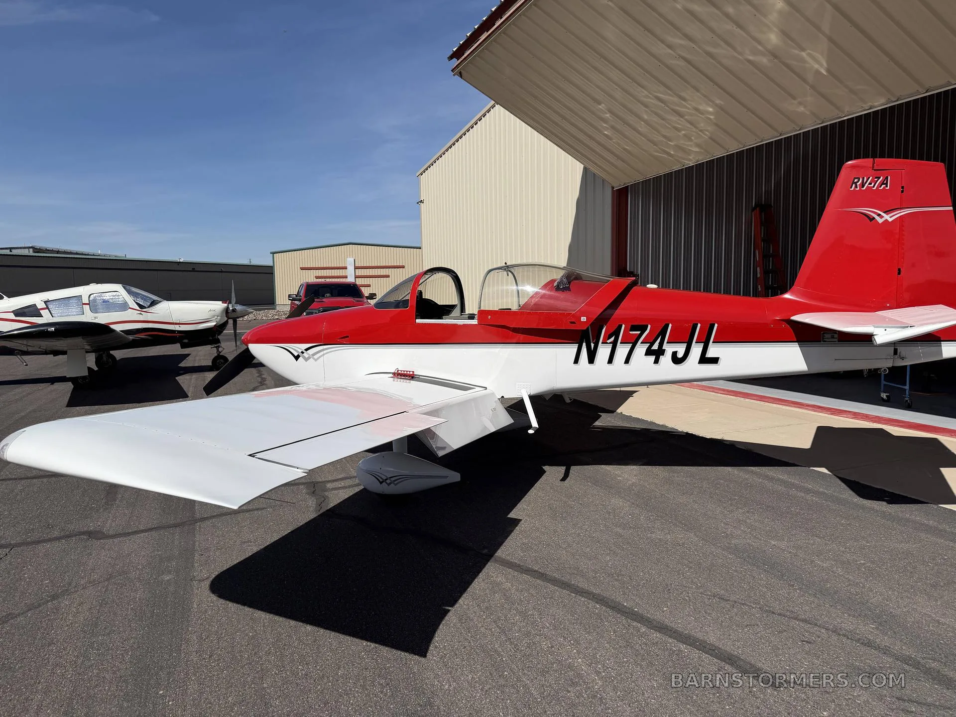 A red and white Vans RV-7A aircraft, registration N174JL, is parked on the tarmac next to another aircraft, viewed from the side.