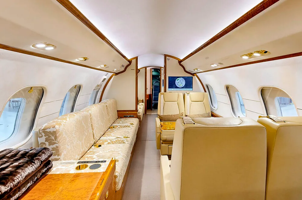 A view of the business jet's cabin interior, showing a long patterned couch, beige seats, and a large display screen.