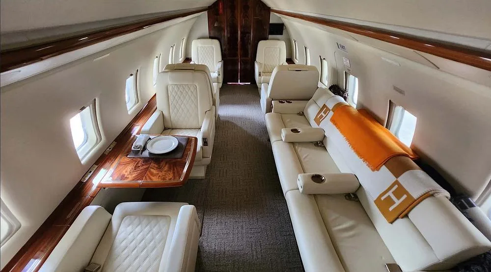 The luxurious interior cabin of a private jet features cream leather seats, a wooden table, and an orange blanket.