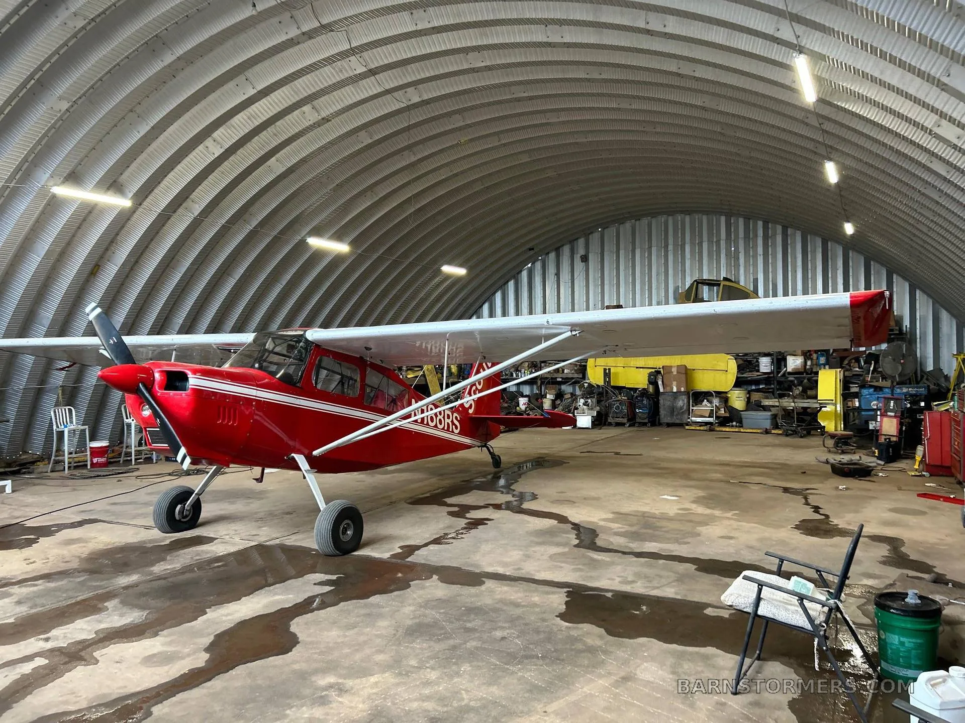 A red Scout N188RS aircraft with white stripes is parked inside a metal hangar, viewed from the front-left.