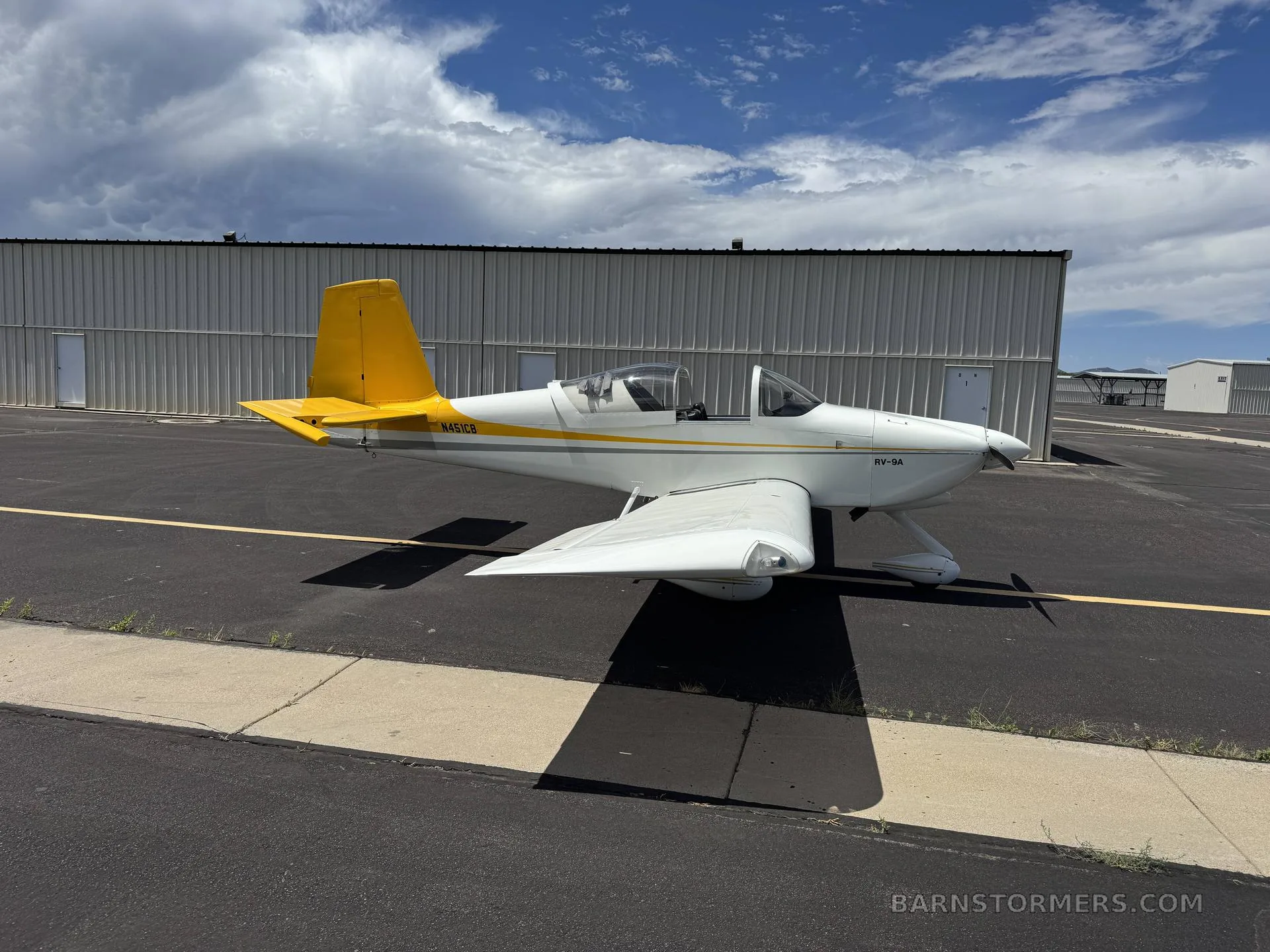 A white RV-9A aircraft with a yellow tail and stripe, parked on an asphalt ramp next to a hangar under a partly cloudy sky.