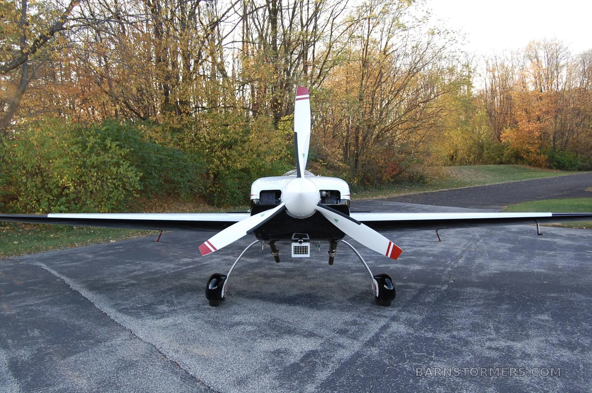 A white, black, and red Staudacher 300 aerobatic aircraft is parked on an asphalt surface with autumn trees in the background, viewed from the front.