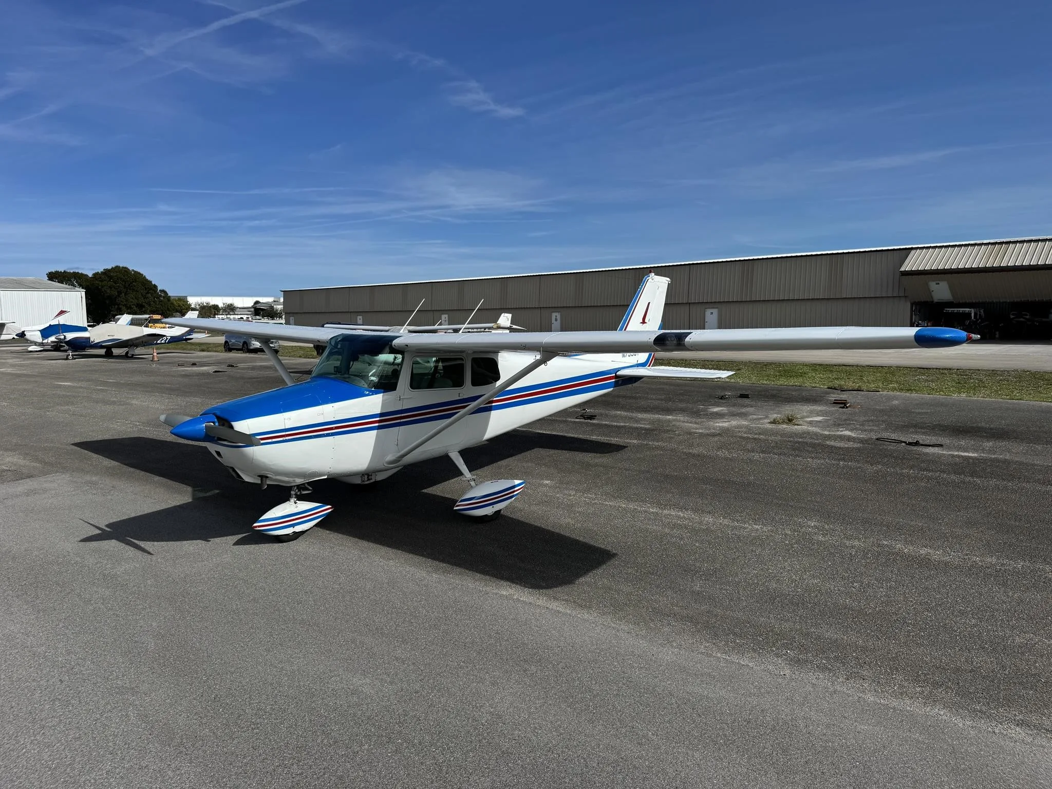 A white and blue single-engine propeller aircraft is parked on an asphalt tarmac with hangars in the background under a blue sky.