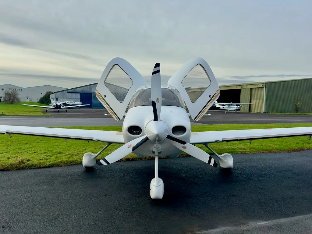 A frontal view of a white Cirrus SR22 aircraft with N322CW registration and open gull-wing doors, parked on a tarmac.