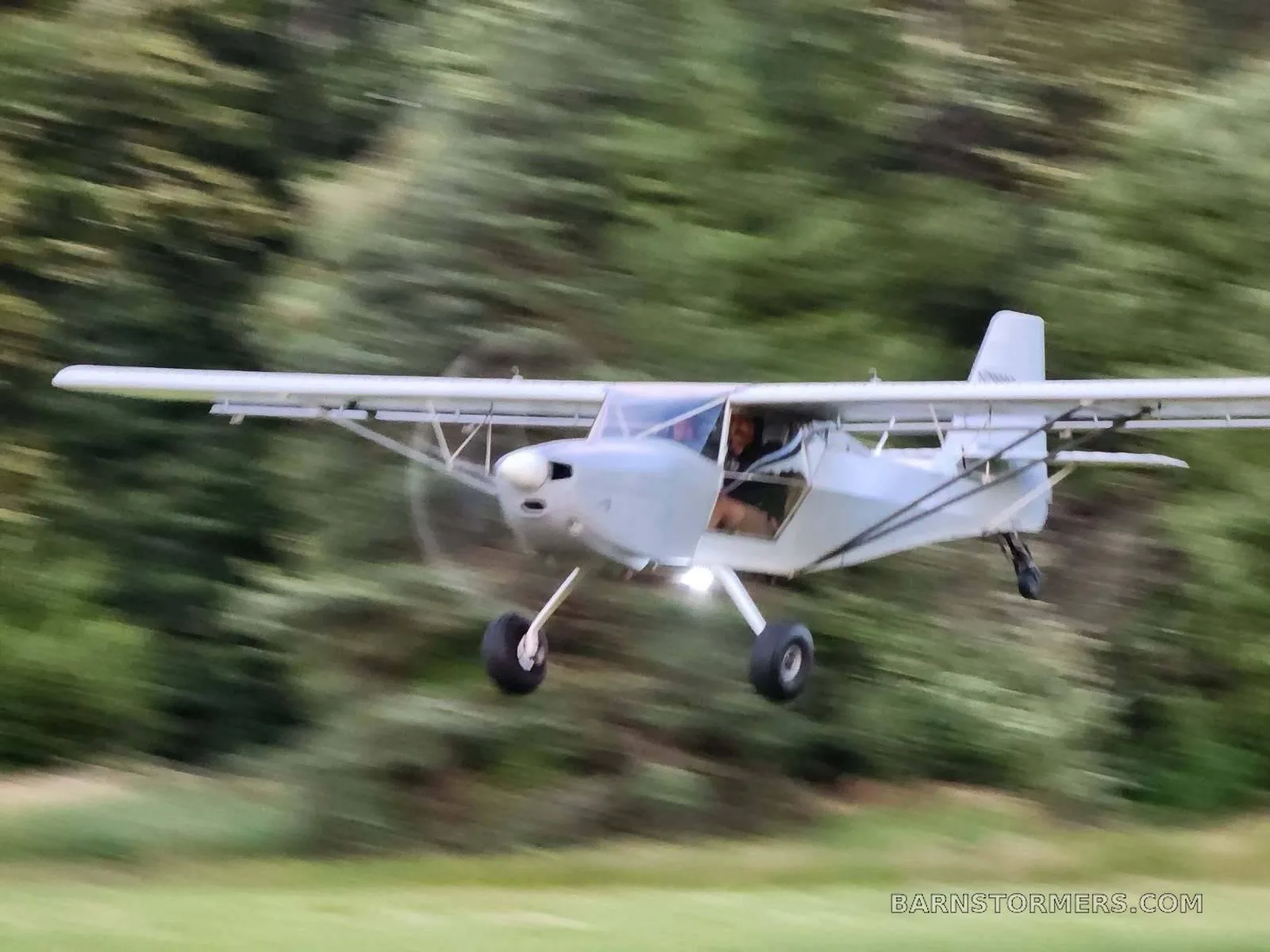 A light grey single-engine aircraft with a high wing and large tires is captured in flight, with a green forest background blurred by motion.