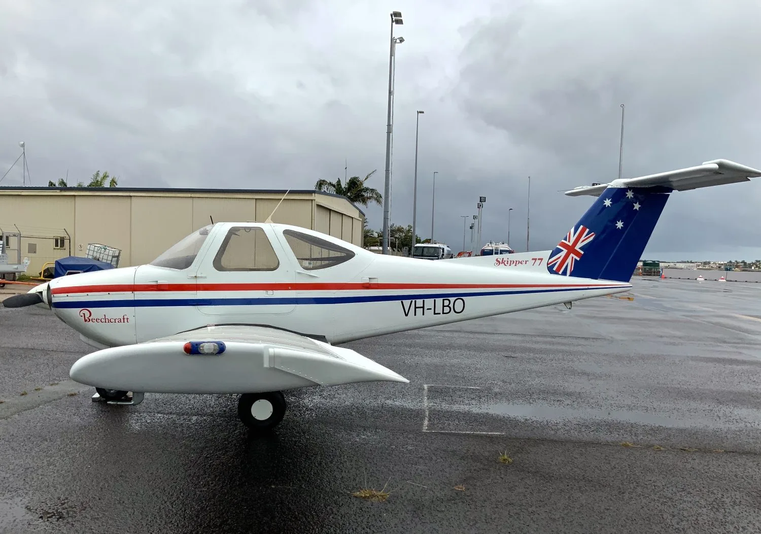 A white Beechcraft Skipper 77 with red and blue stripes and Australian flag tail, registration VH-LBO, parked on a wet tarmac under a cloudy sky.