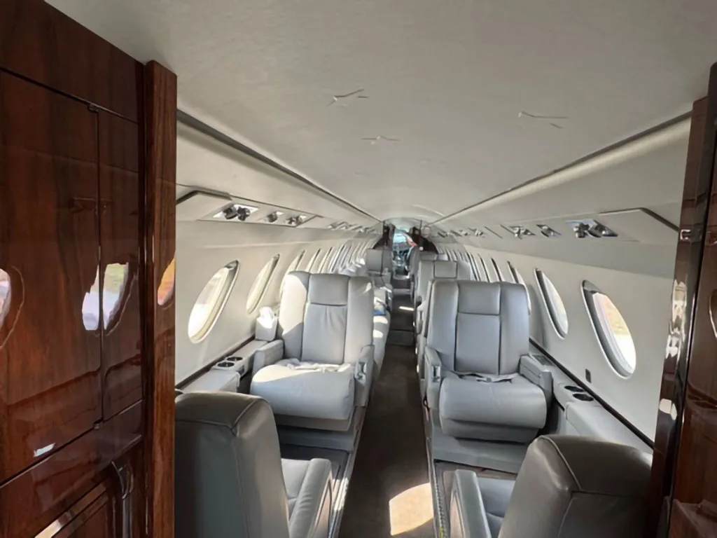 View of a business jet cabin with light gray leather seats and polished wood cabinetry, looking towards the rear.