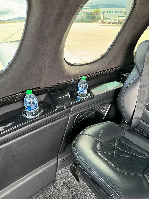 A close-up of the aircraft's black leather interior, showing a seat, cup holders with water bottles, and a window view of the tarmac.