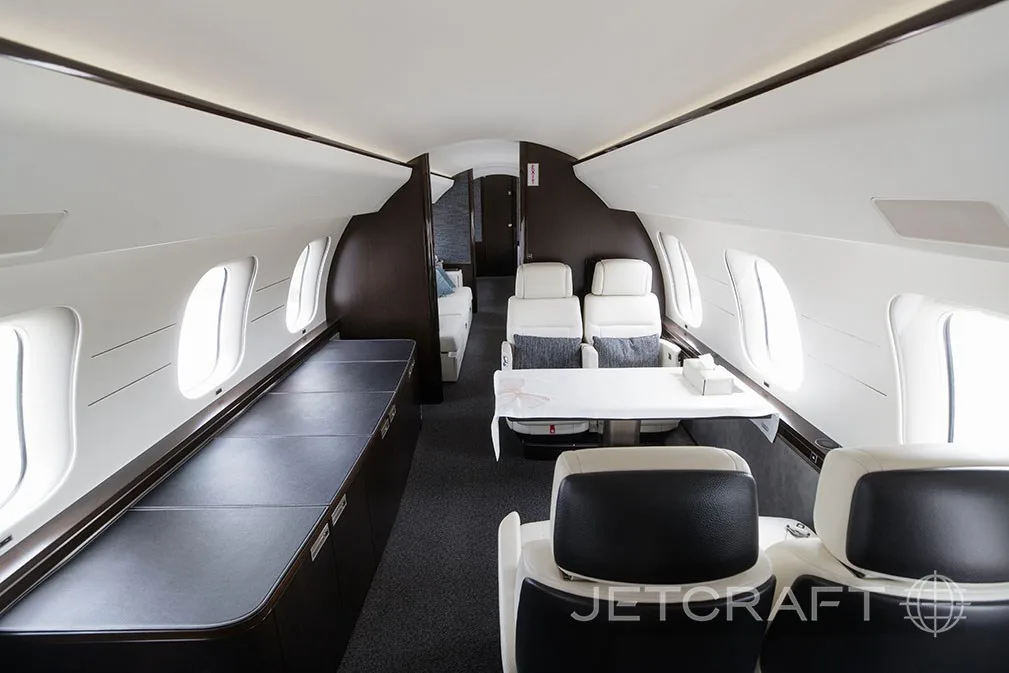 A private jet cabin showing a dining area with a white tablecloth and a lounge area in the background.