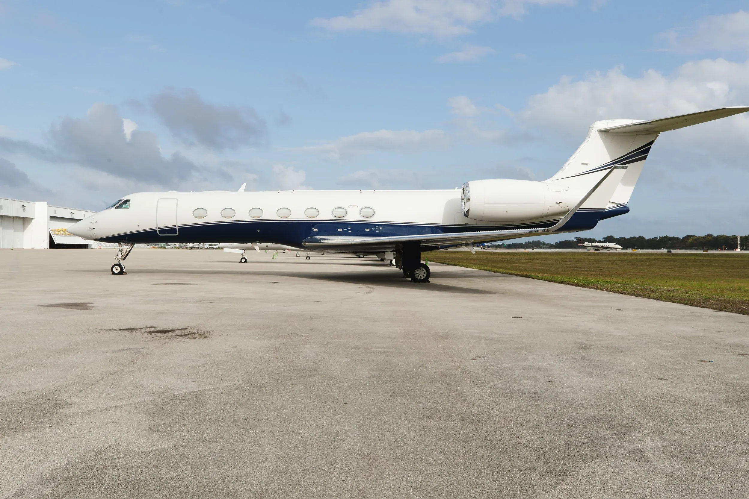 A white and blue private jet, likely a Gulfstream, is parked on an airport tarmac with hangars in the background.