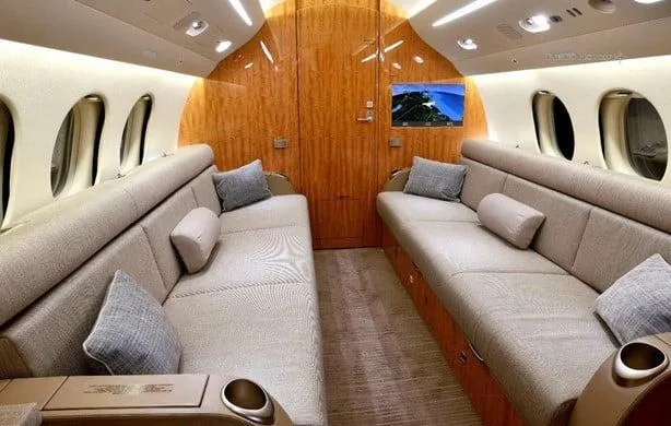 A private jet cabin interior with two beige couches, throw pillows, and a large screen displaying a landscape image on the wooden bulkhead.