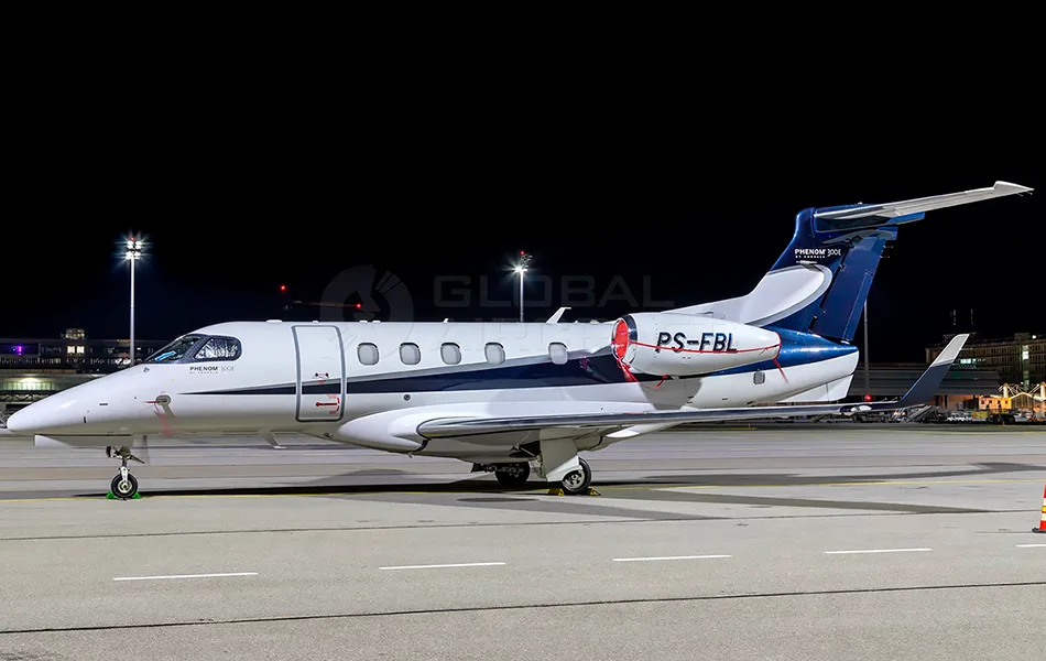 A white and blue Embraer Phenom 300E business jet with registration PS-FBL is parked on an airport tarmac at night.