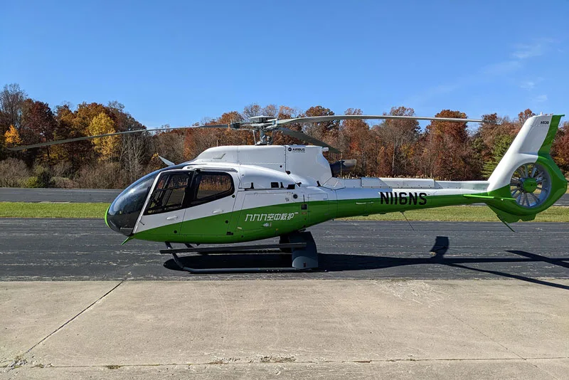 A white and green Airbus H130 helicopter with registration N116NS sits on a tarmac with autumn trees in the background.