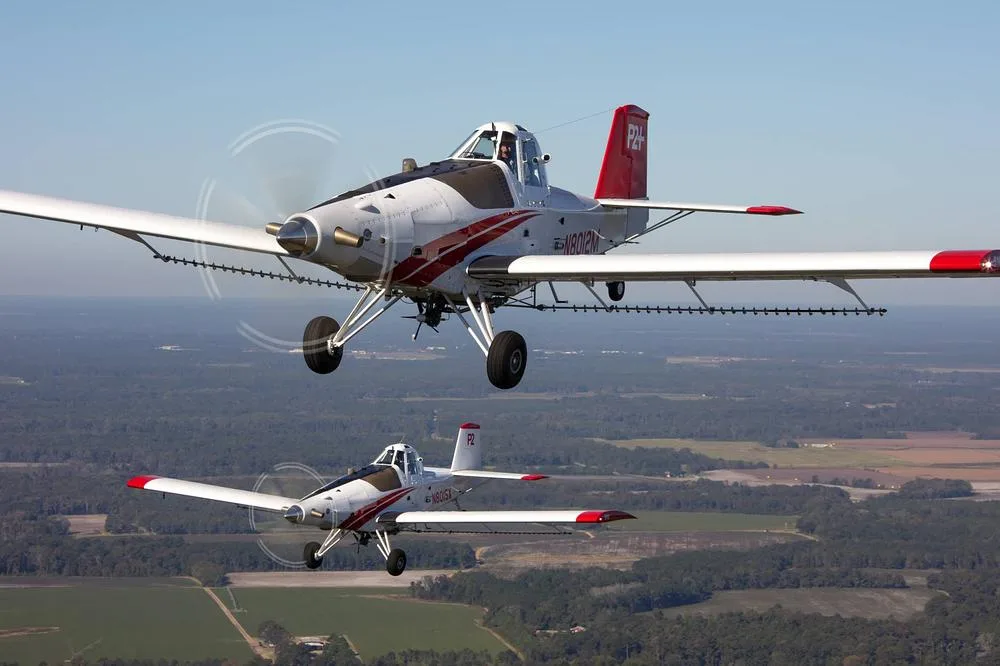 Two white and red crop duster aircraft flying in formation over a forested and agricultural landscape.