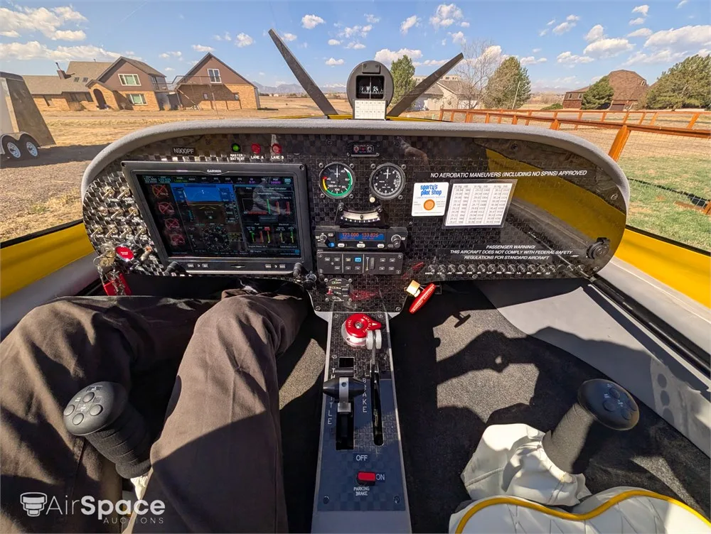 The cockpit of a yellow aircraft shows a modern glass panel display, traditional gauges, and flight controls, with a view of houses and trees outside.
