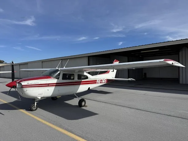 A white and red Cessna 210 Centurion aircraft, N87MS, is parked on an asphalt tarmac next to an open hangar under a blue sky.