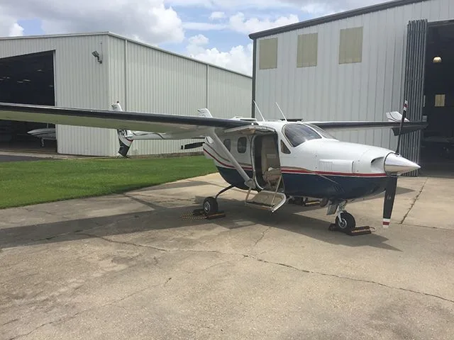 A white and blue single-engine aircraft with an open door, parked on a concrete apron in front of hangars.