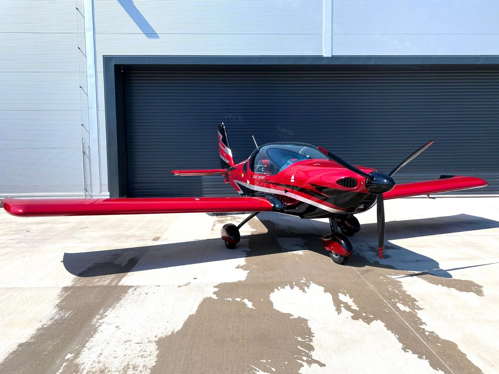 A red and black Viper SD4 light sport aircraft with its canopy closed, viewed from the front, parked in front of a hangar.