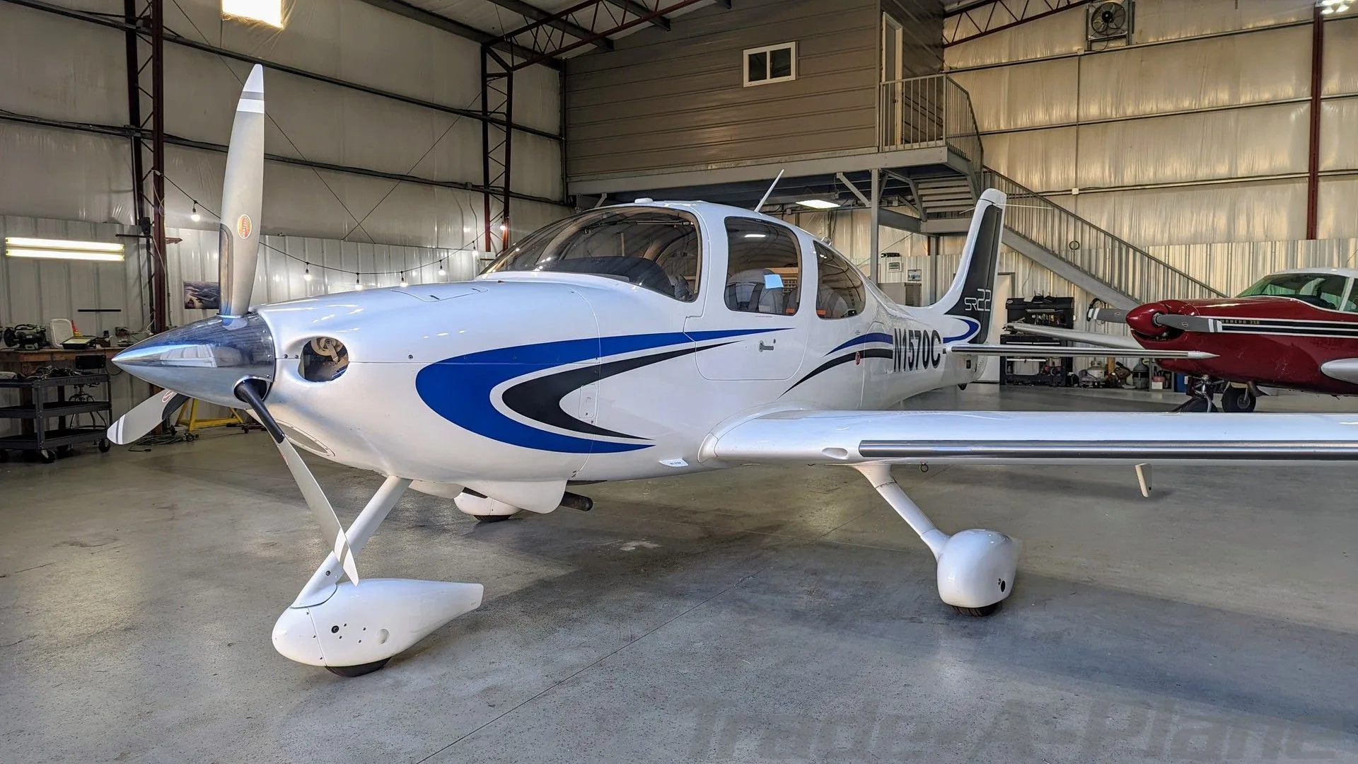 Front view of a white Cirrus SR22 aircraft with blue and black stripes, registration N1570C, parked inside a hangar.