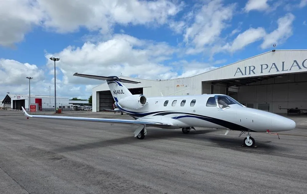 A white Cessna Citation M2 jet with blue and black stripes, tail number N540JL, parked on a tarmac with a hangar in the background.