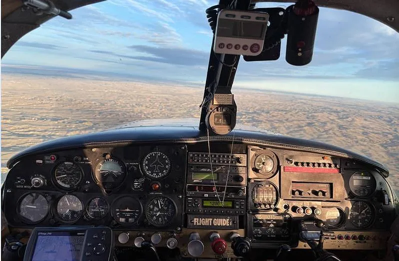 The instrument panel of a small aircraft is shown with various gauges and avionics, looking out over a landscape at sunset.
