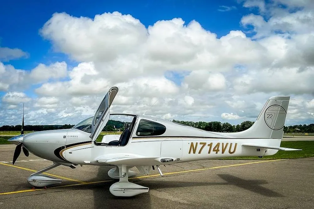 A white and silver Cirrus SR22 aircraft with gold and black accents, registration N714VU, parked on an airfield with its door open under a blue sky with clouds.