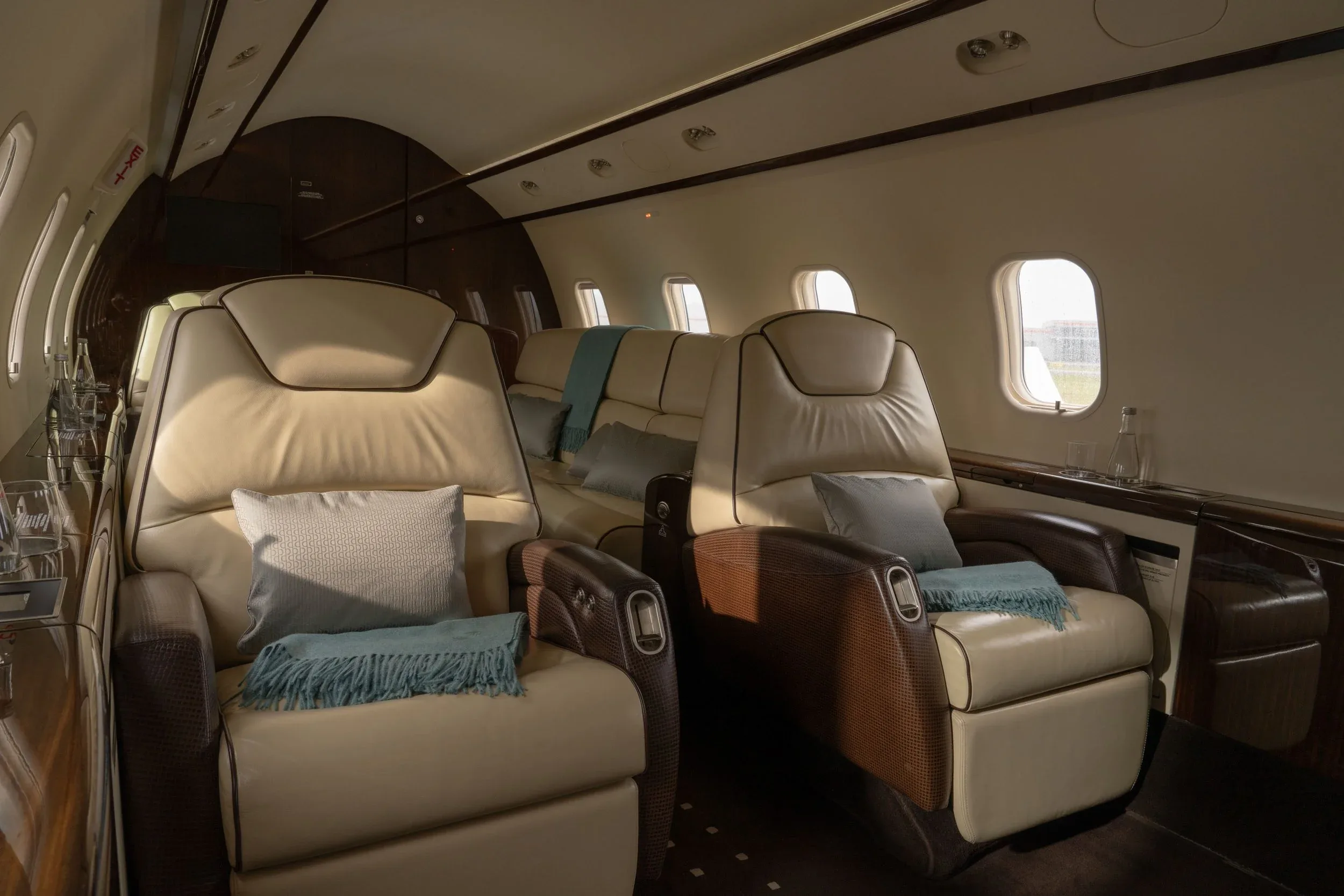 A side view of the aircraft cabin highlights two plush beige leather seats with teal throws and pillows, next to large windows.