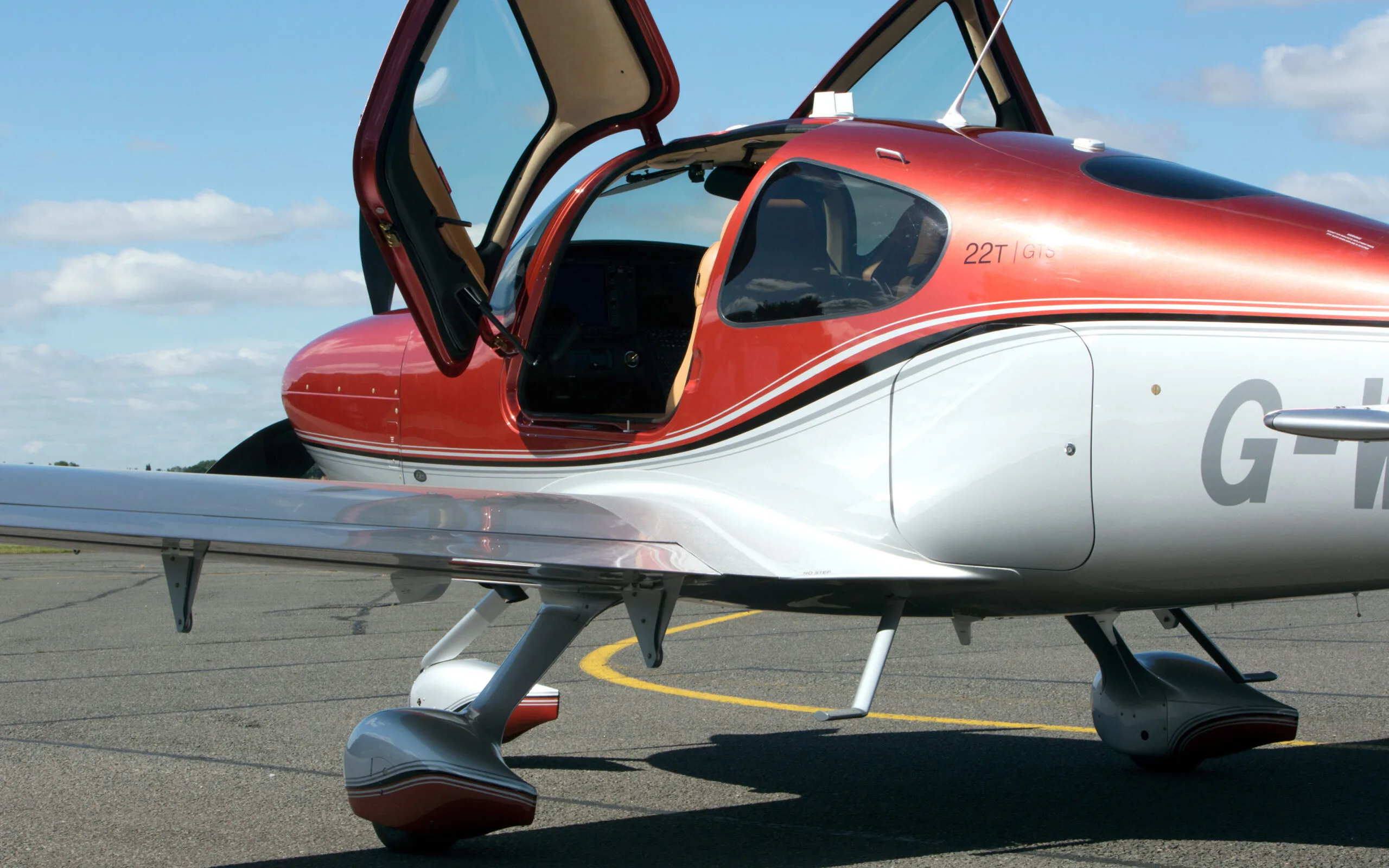 A close-up side view of a red and silver Cirrus SR22T GTS aircraft with its door open, parked on an airfield.
