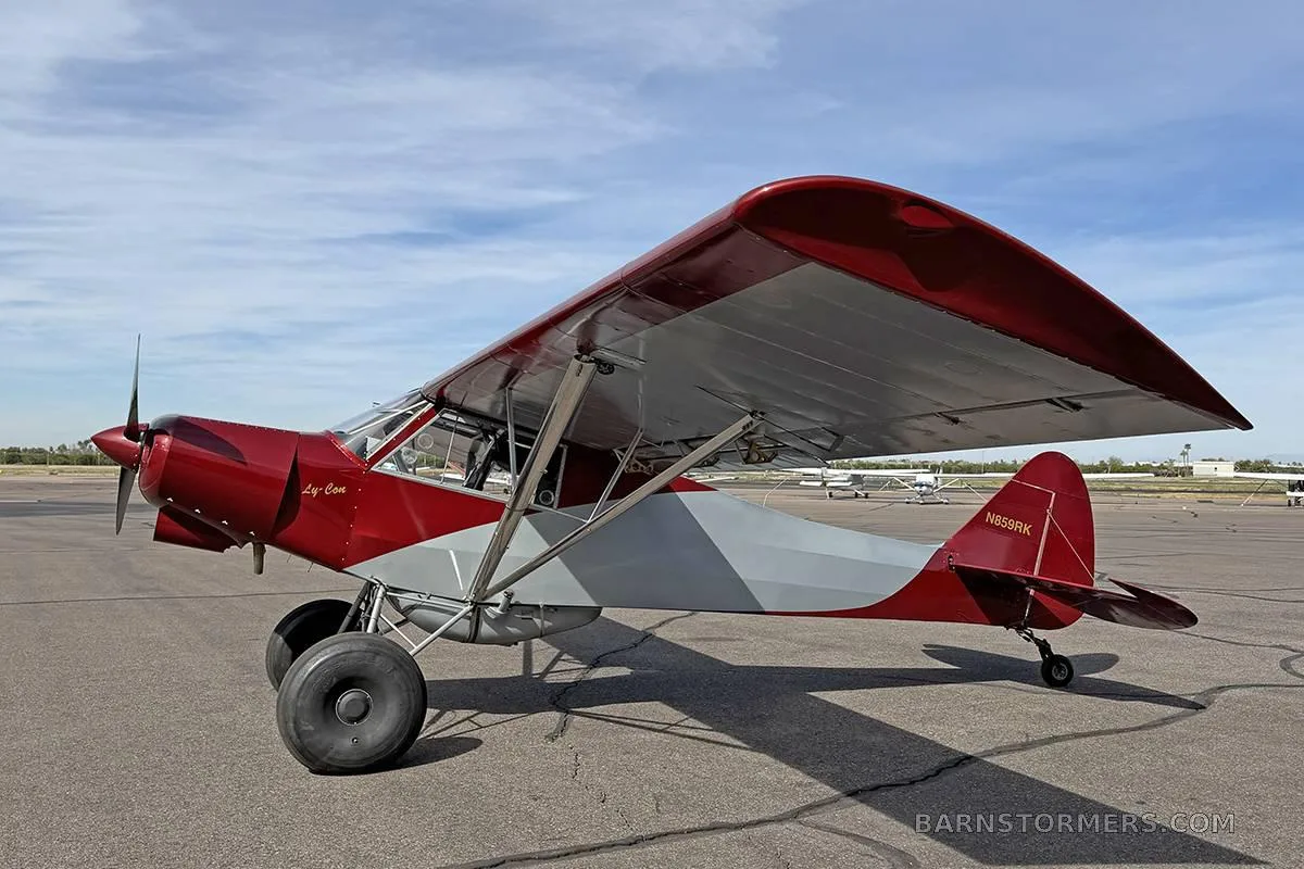 A red and grey high-wing aircraft with large tires, seen from the right side on an airfield.