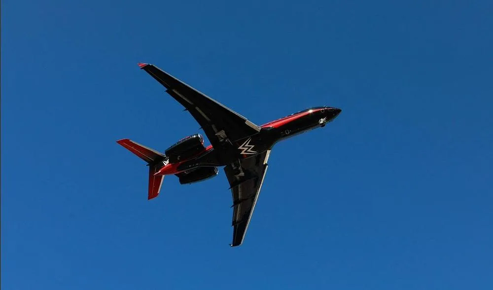 A black and red Bombardier Global 7500 business jet with a white logo on its tail and underside flies against a clear blue sky.