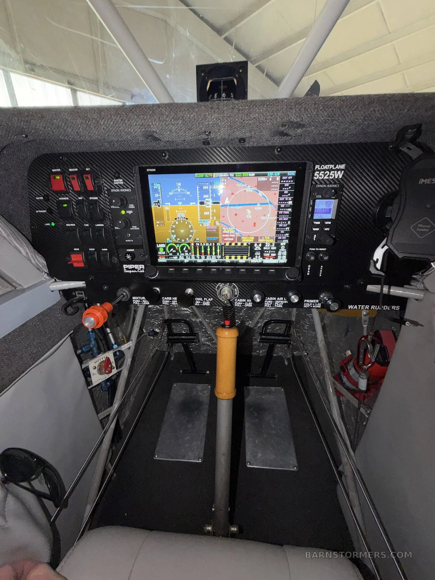 The cockpit of a seaplane features a large digital display, various gauges, switches, and rudder pedals.