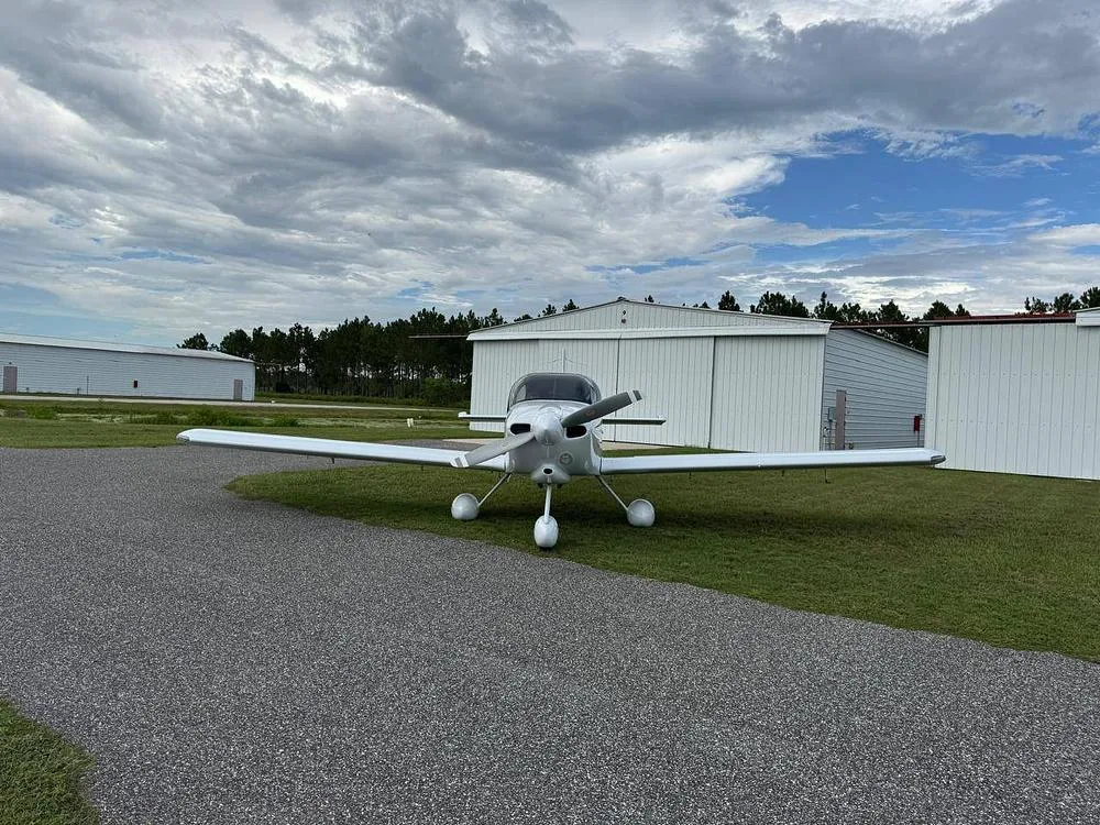 A front view of a white Van's RV-10 aircraft, registration N511DL, parked on grass next to a gravel path and hangars.