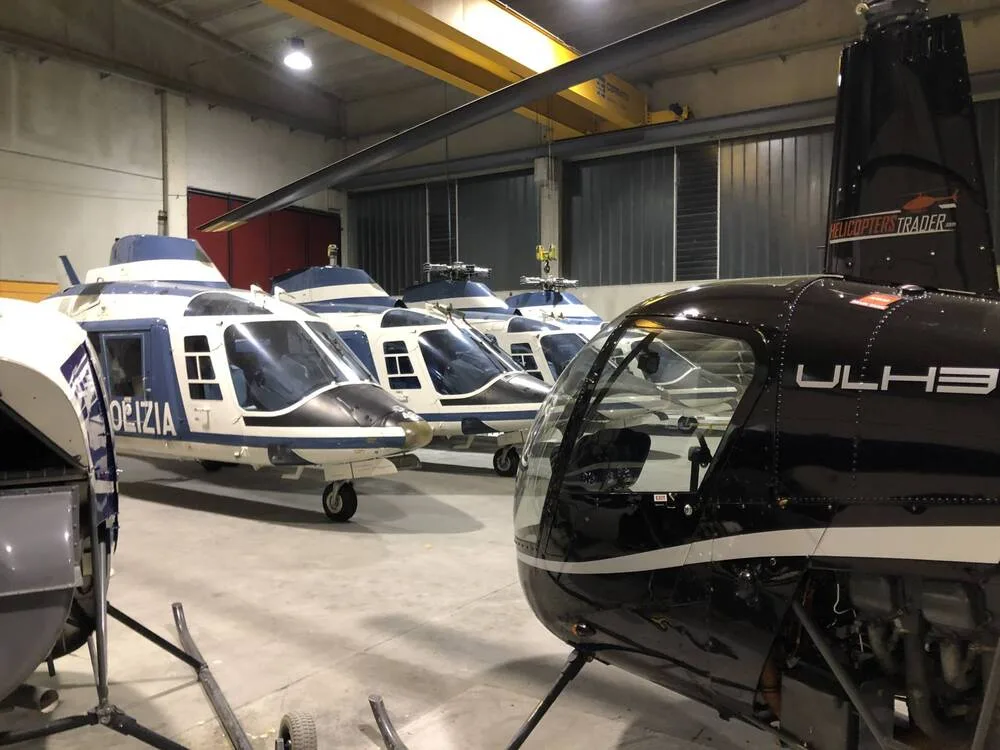 Multiple helicopters, some marked "POLIZIA", are parked in a hangar, with one in the foreground showing "HELICOPTERS TRADER" on its tail.