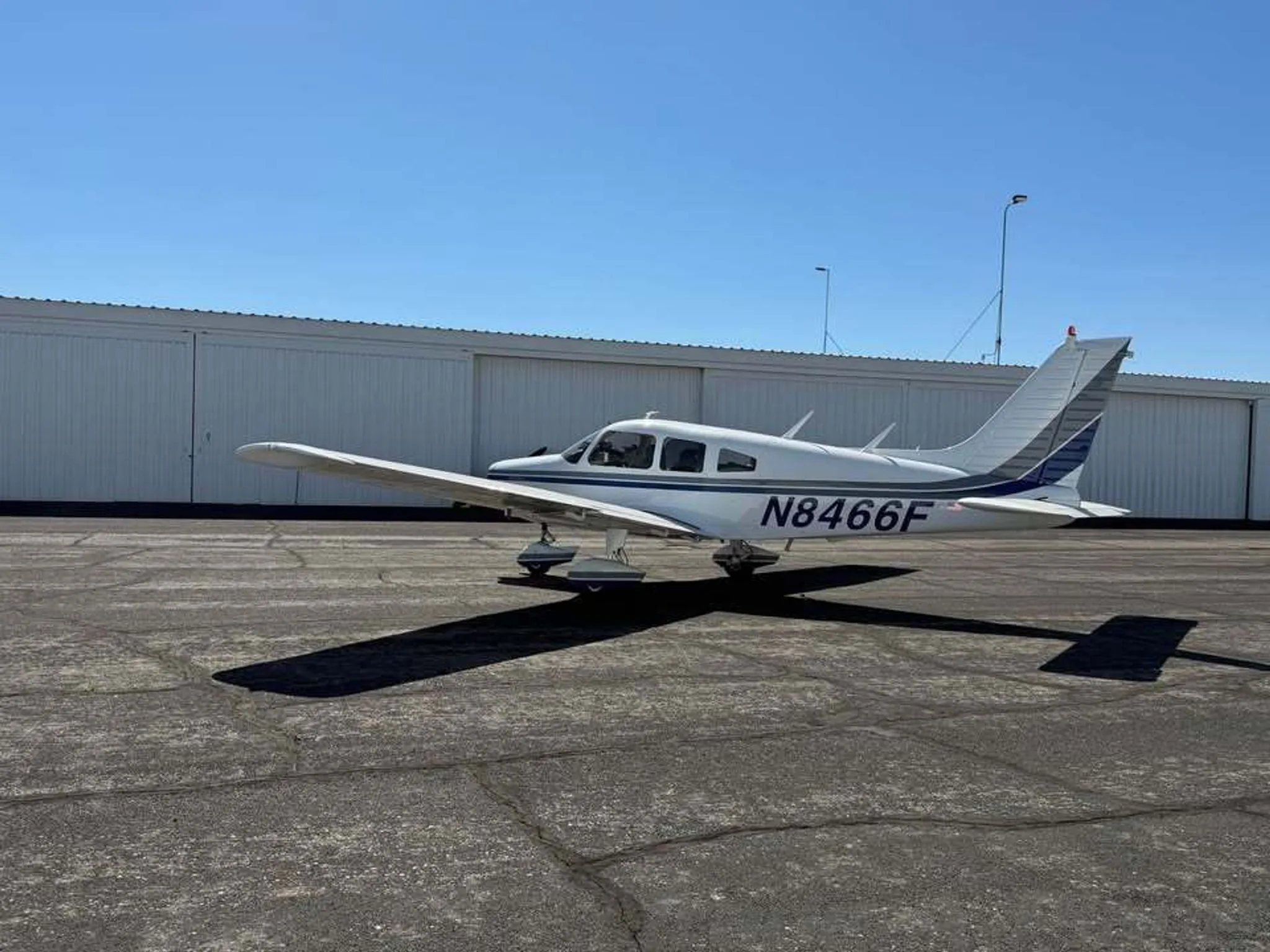 A white Piper Warrior II aircraft with blue and gray stripes and registration N8466F parked on an asphalt apron.