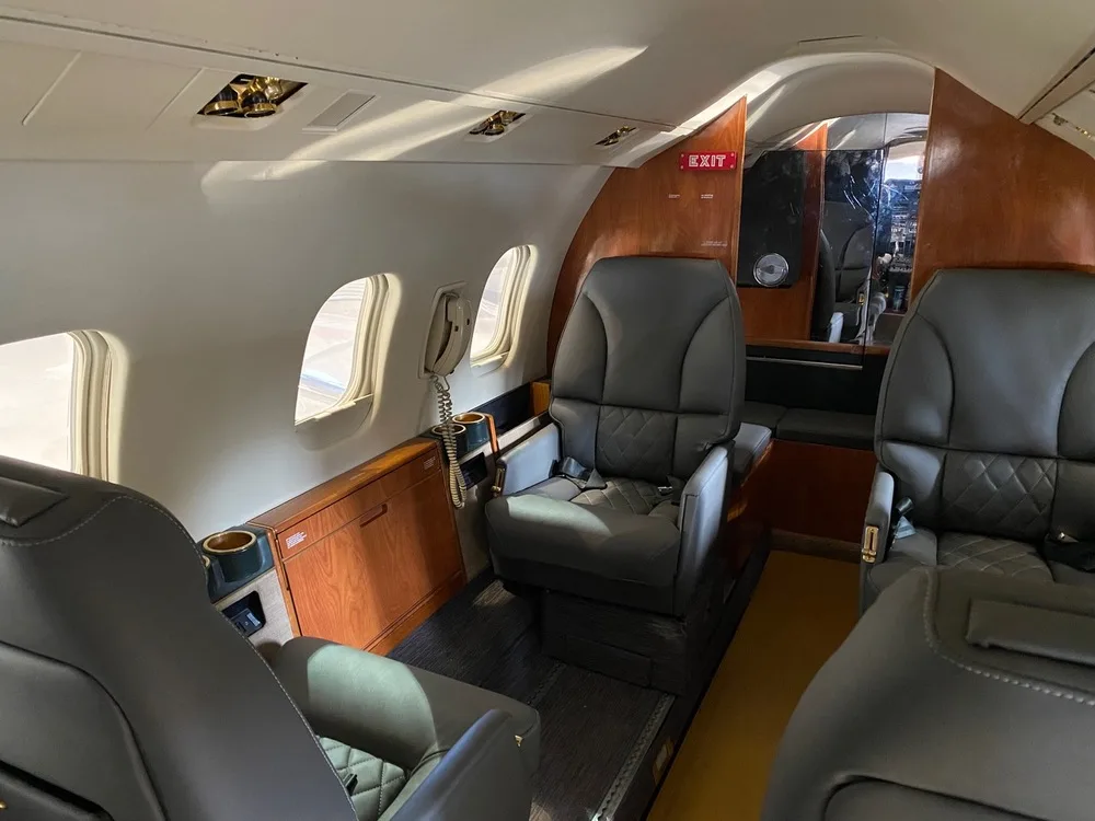 The interior cabin of a business jet, featuring dark grey leather seats, wood paneling, and an 'EXIT' sign.