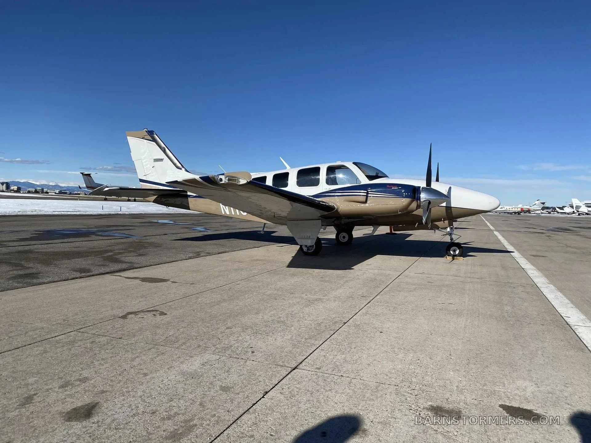 A white, blue, and tan twin-engine aircraft is parked on an asphalt runway under a clear blue sky, with other aircraft in the distance.