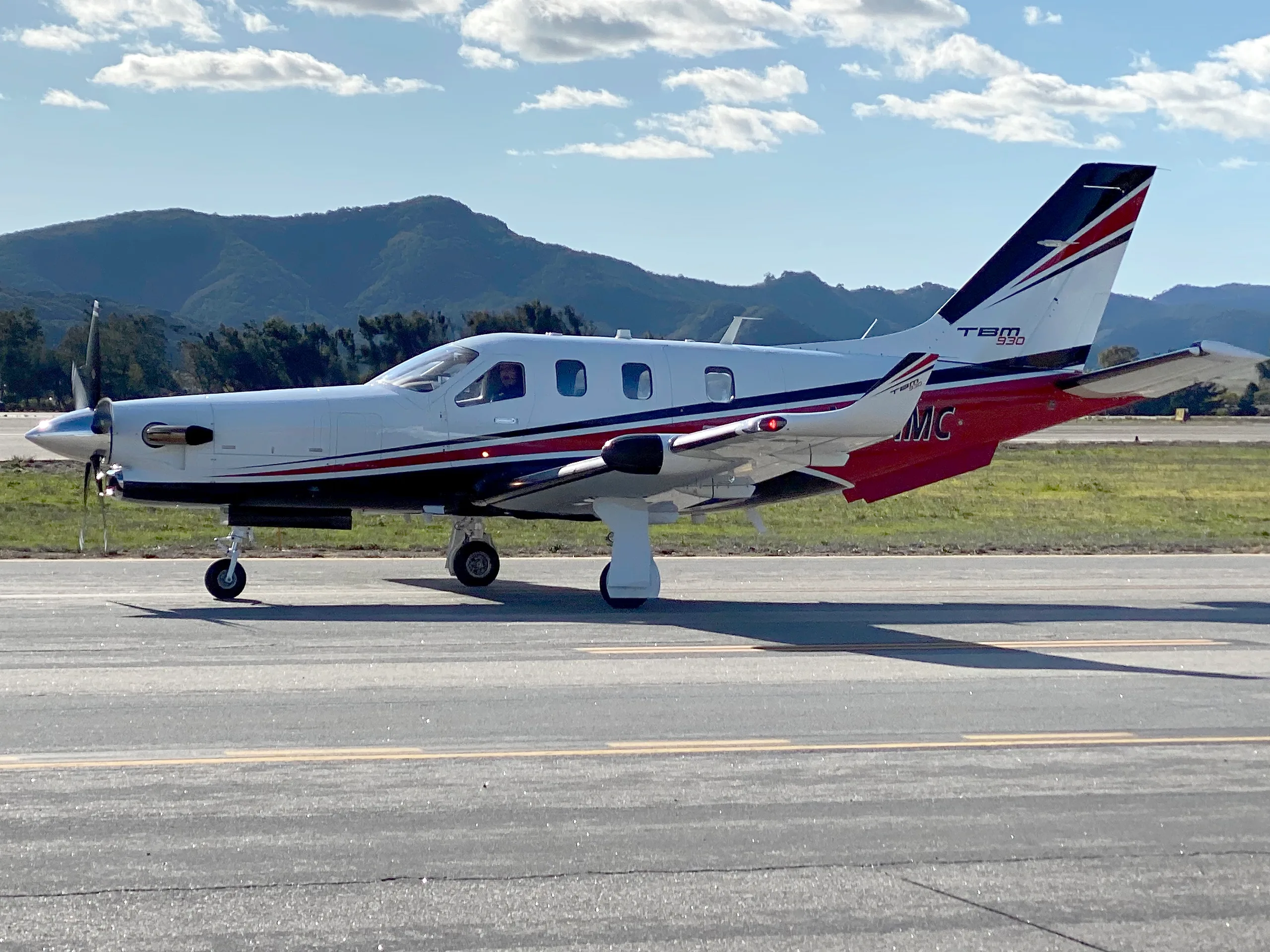 A white Daher TBM 930 aircraft with red and black stripes is seen from the side on an airfield, with mountains in the background.