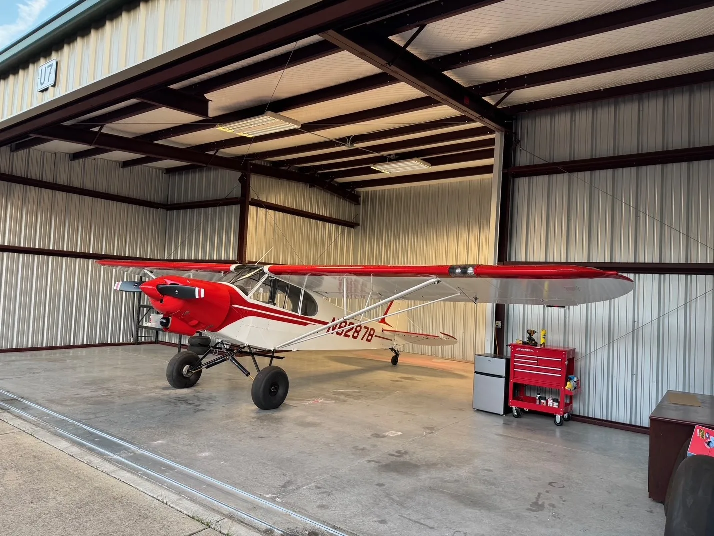 A red and white bush plane, N8287B, with large tires, parked inside a hangar.