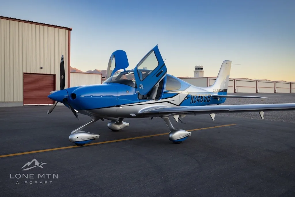 A blue and silver Cirrus SR22 aircraft with open doors parked on an airfield at sunset, with an air traffic control tower in the background.