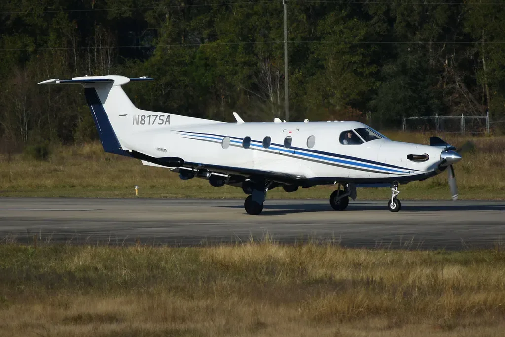 A white Pilatus PC-12 turboprop aircraft with blue and black stripes taxis on an airfield.