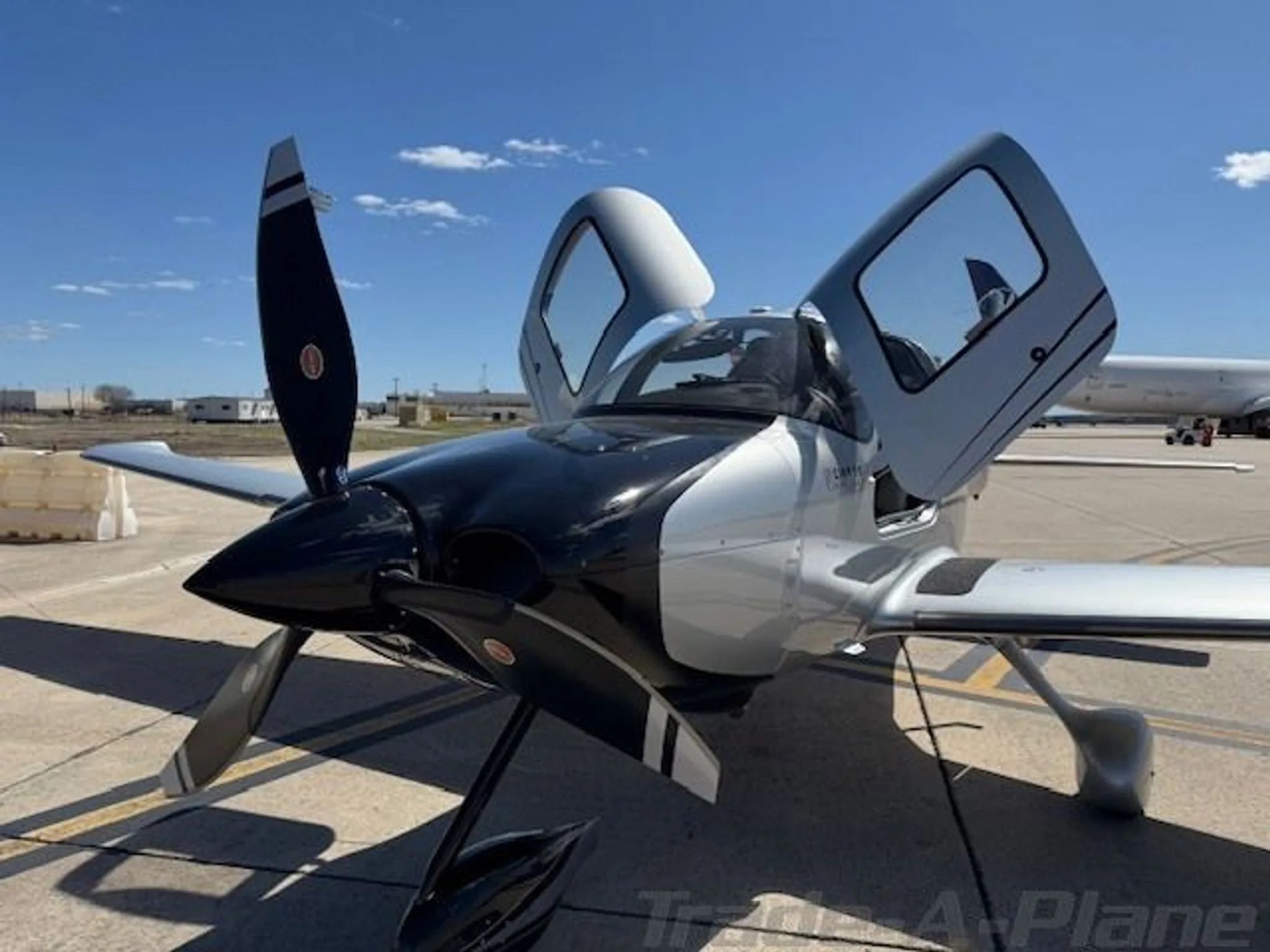 A black and silver Cirrus SR22 GTS Turbo aircraft with open doors is parked on a tarmac under a blue sky.