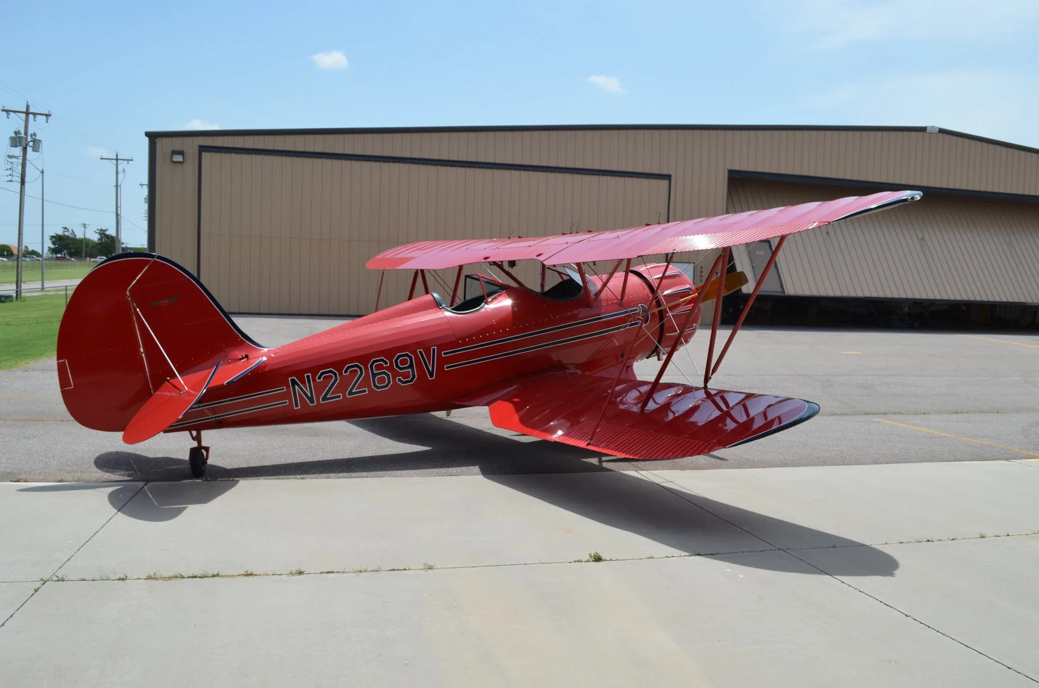 A full side view of the red biplane N2269V parked on an asphalt apron next to a tan hangar.