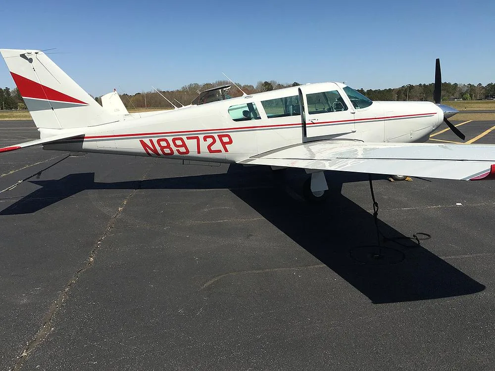 A side view of the white Piper Cherokee 180 (N8972P) with red and grey stripes parked on an asphalt ramp under a clear sky.
