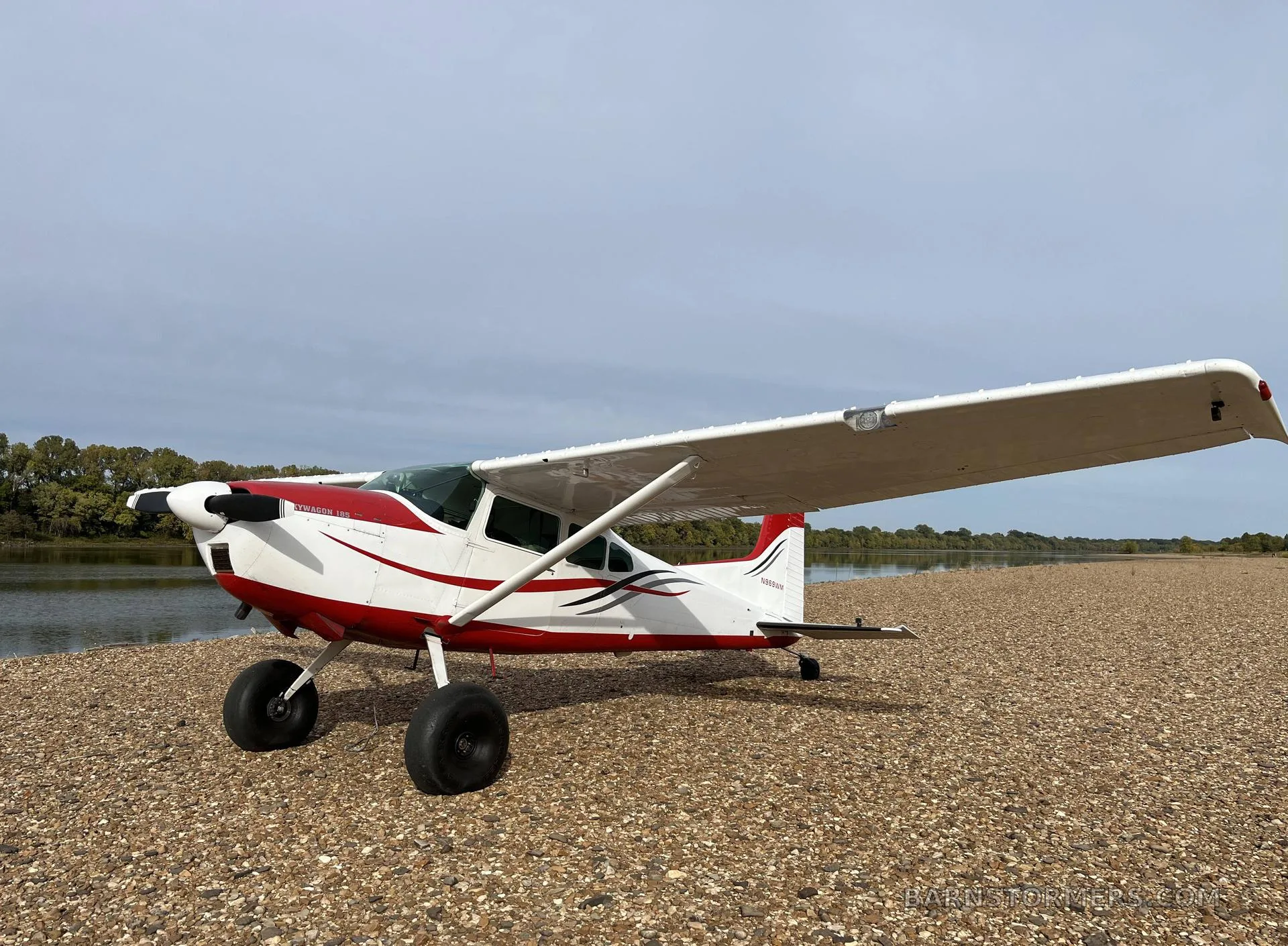 A white and red Cessna 180 aircraft with tundra tires parked on a gravel riverbank under a cloudy sky.