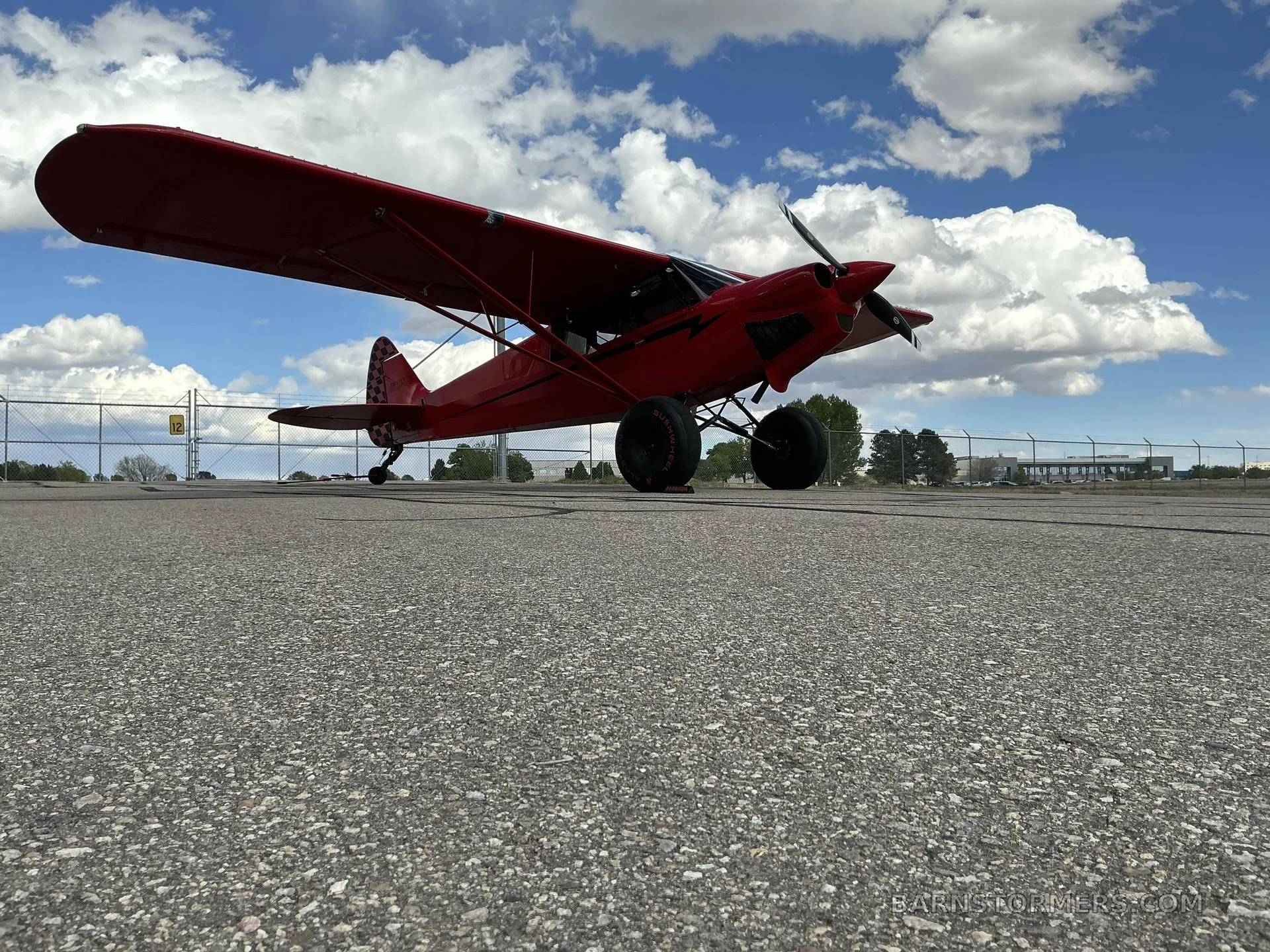 A low-angle view of a red bush plane with large tires parked on an asphalt tarmac under a partly cloudy sky.