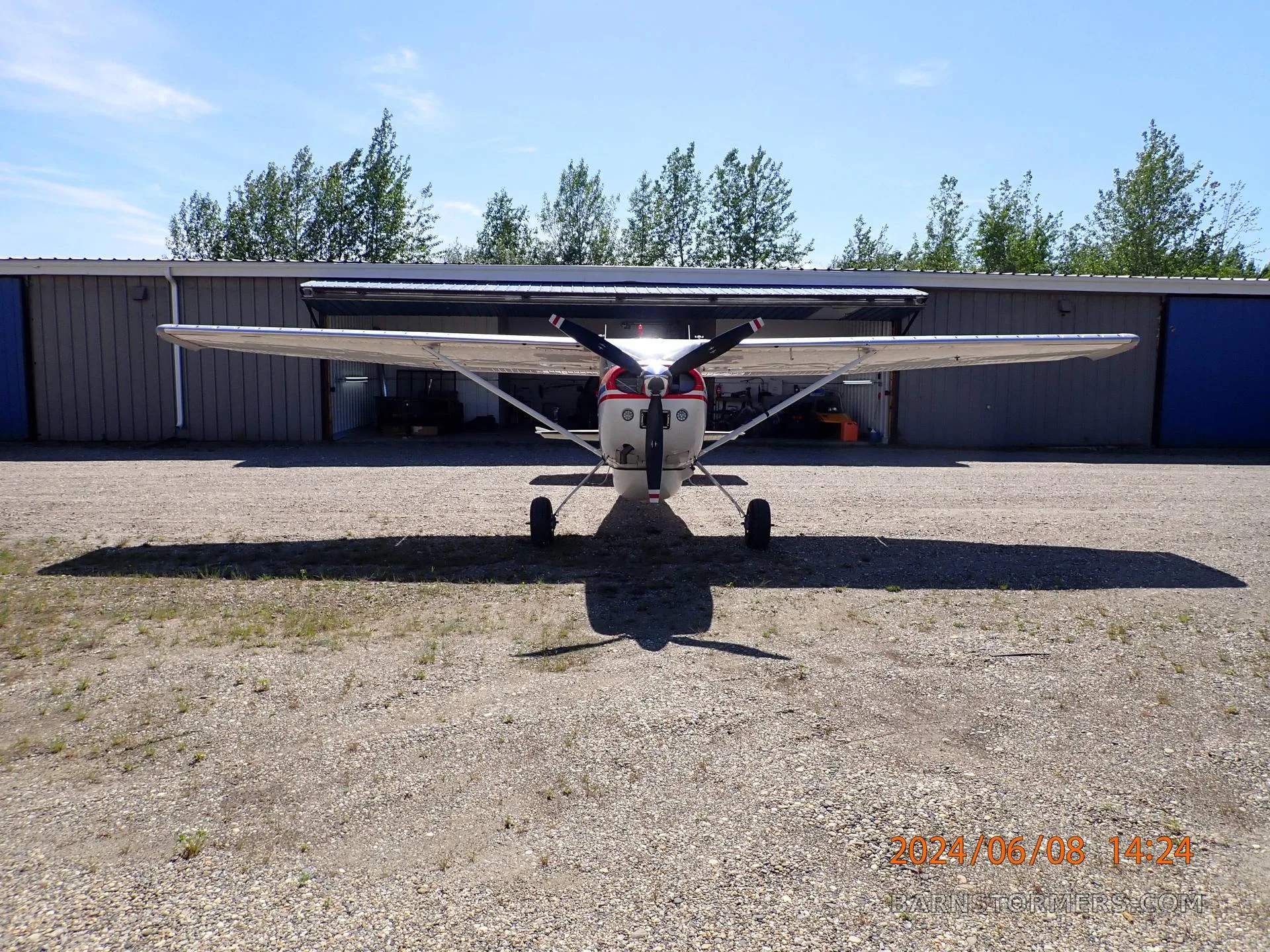 A front view of a red, white, and blue Cessna 180 aircraft parked on a gravel lot in front of hangars.