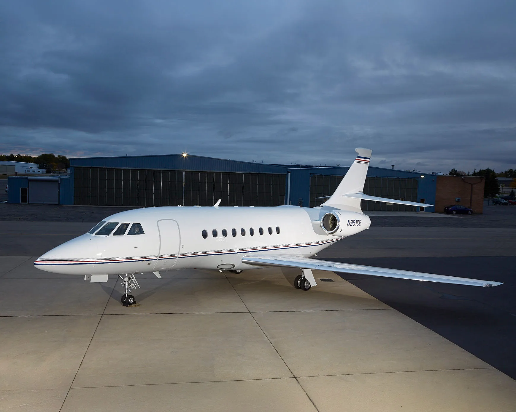 A white Dassault Falcon business jet with blue and red stripes on the tail and fuselage, parked on an tarmac with hangars in the background under a cloudy sky.
