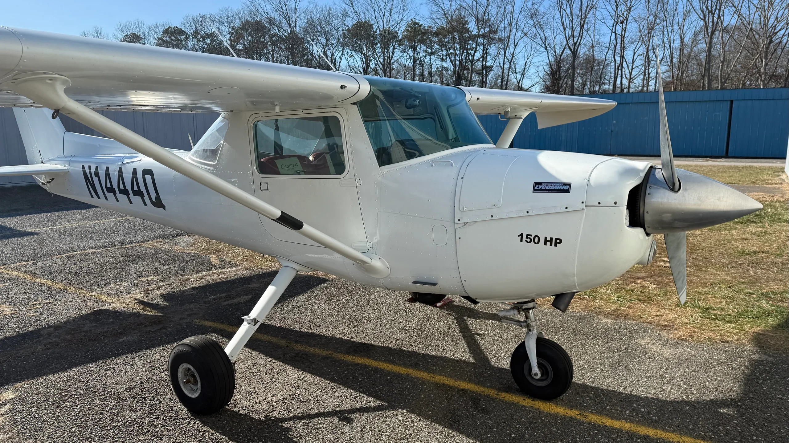 A white Cessna 150 aircraft, registration N1444Q, is parked on an asphalt ramp with a blue hangar in the background.